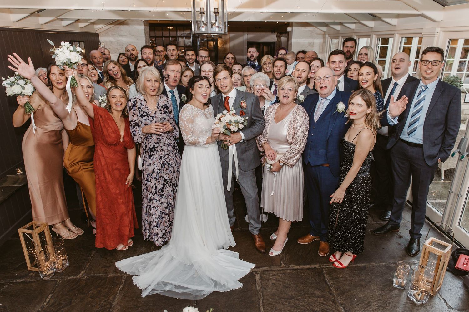 Large group wedding photo with guests in formal attire gathered under elegant lighting.
