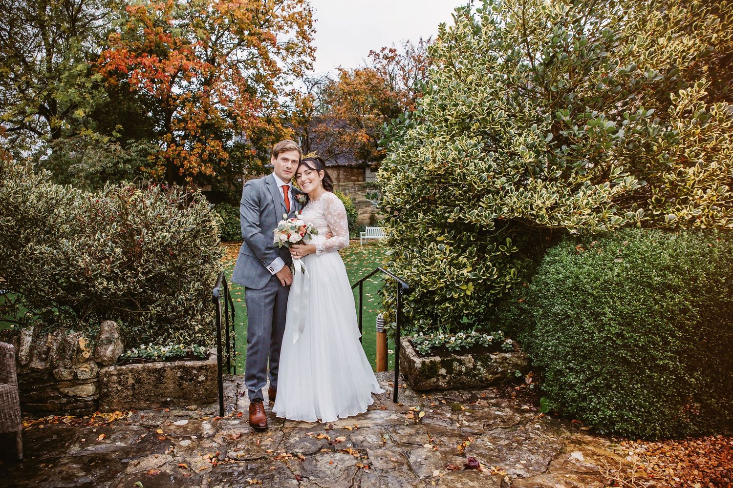 Wedding couple posing on stone steps surrounded by autumn trees and green hedges.