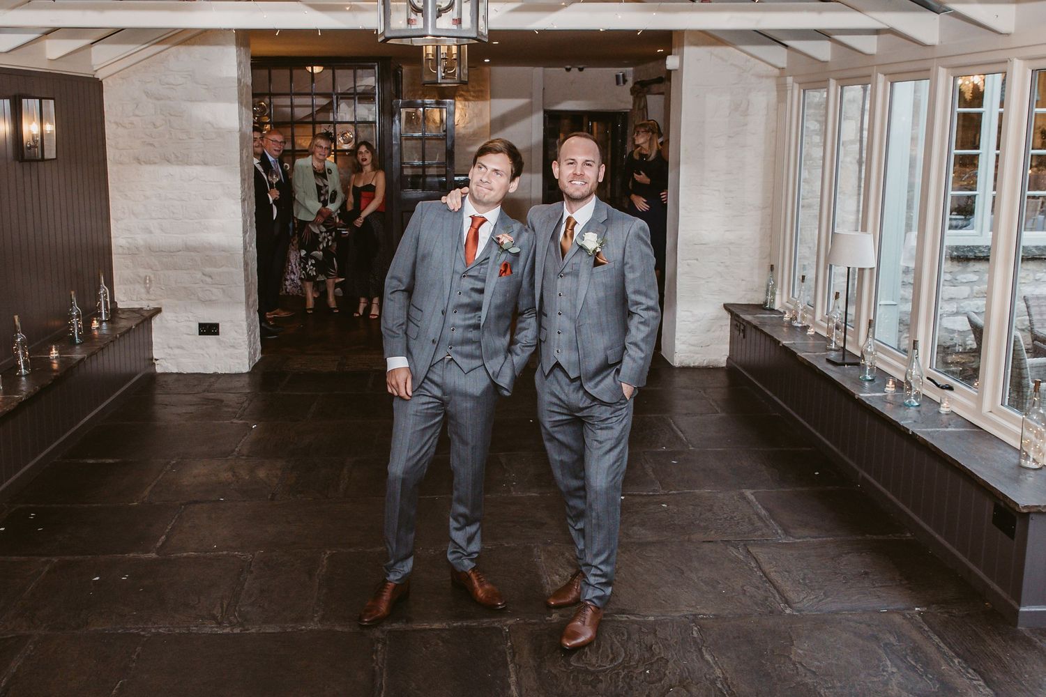 Two groomsmen in matching gray suits and burgundy ties stand together in an elegant indoor venue.