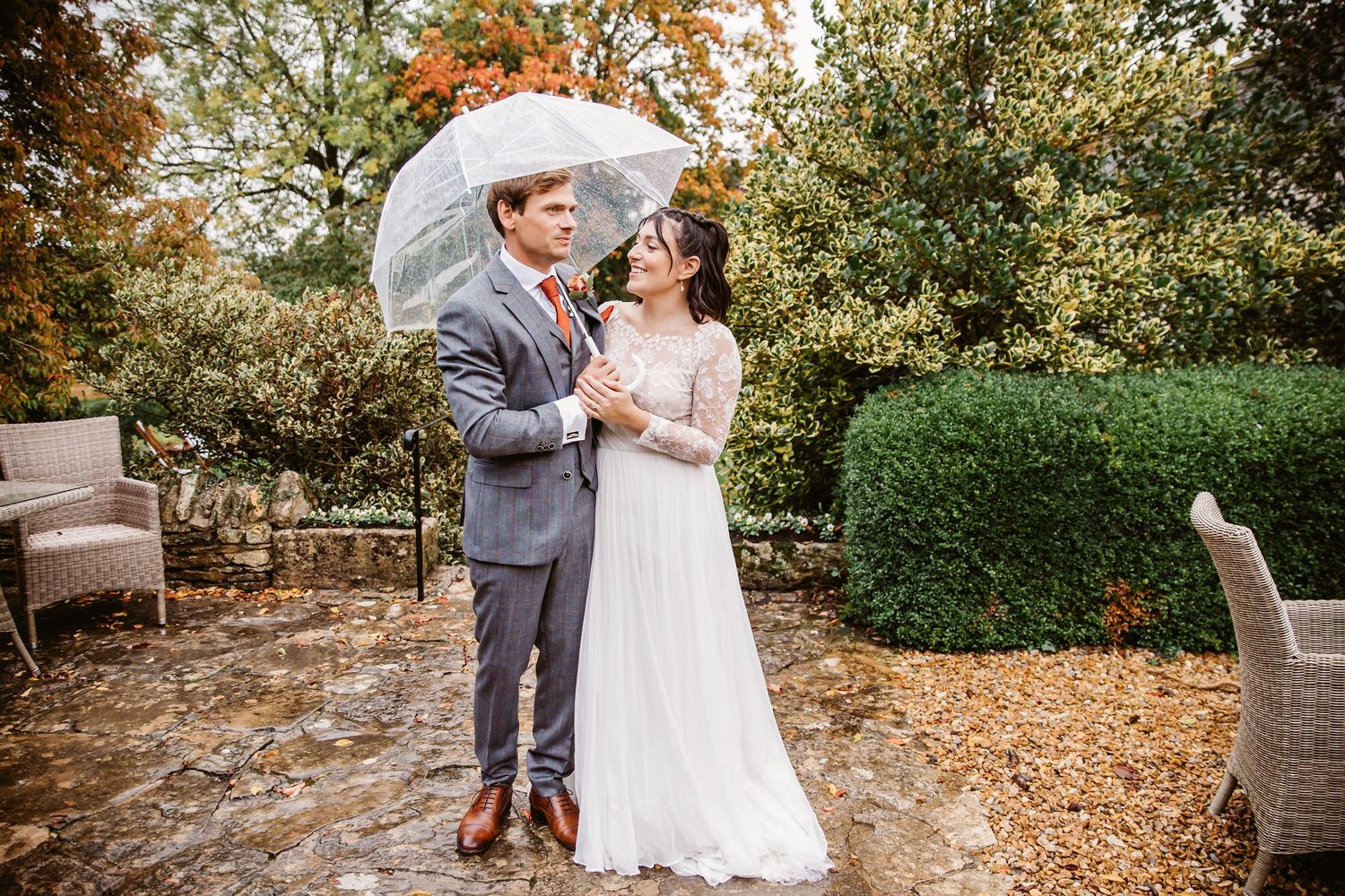 A couple shares a romantic moment under a clear umbrella in a garden setting.