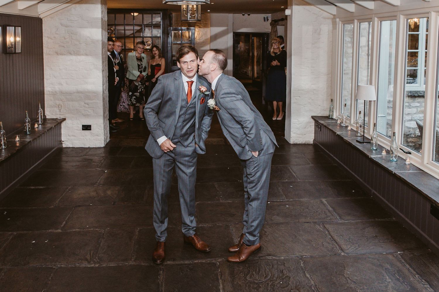 Two groomsmen in matching gray suits share a playful moment at wedding reception.
