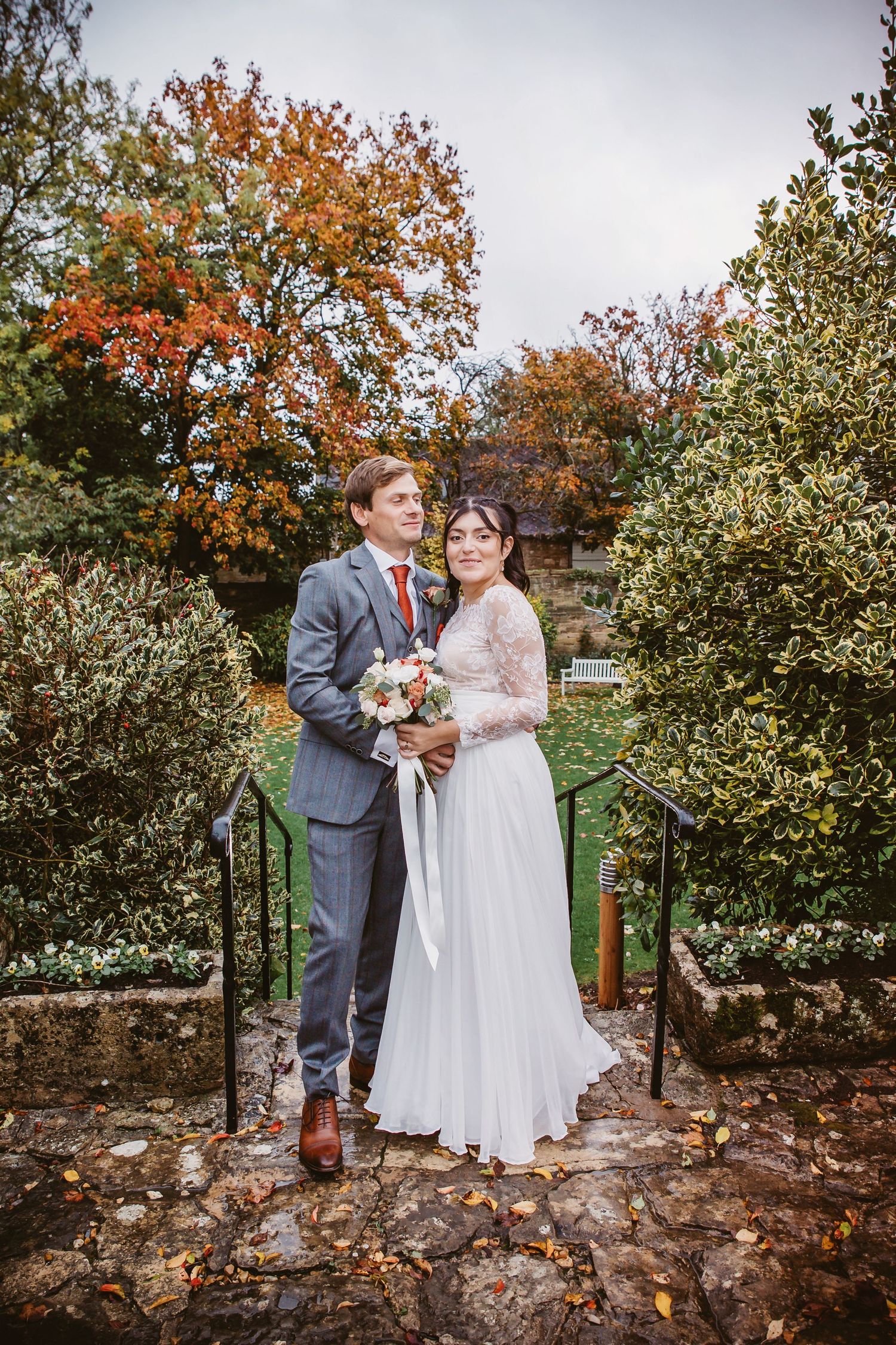Wedding couple standing on stone steps with autumn foliage and garden greenery.