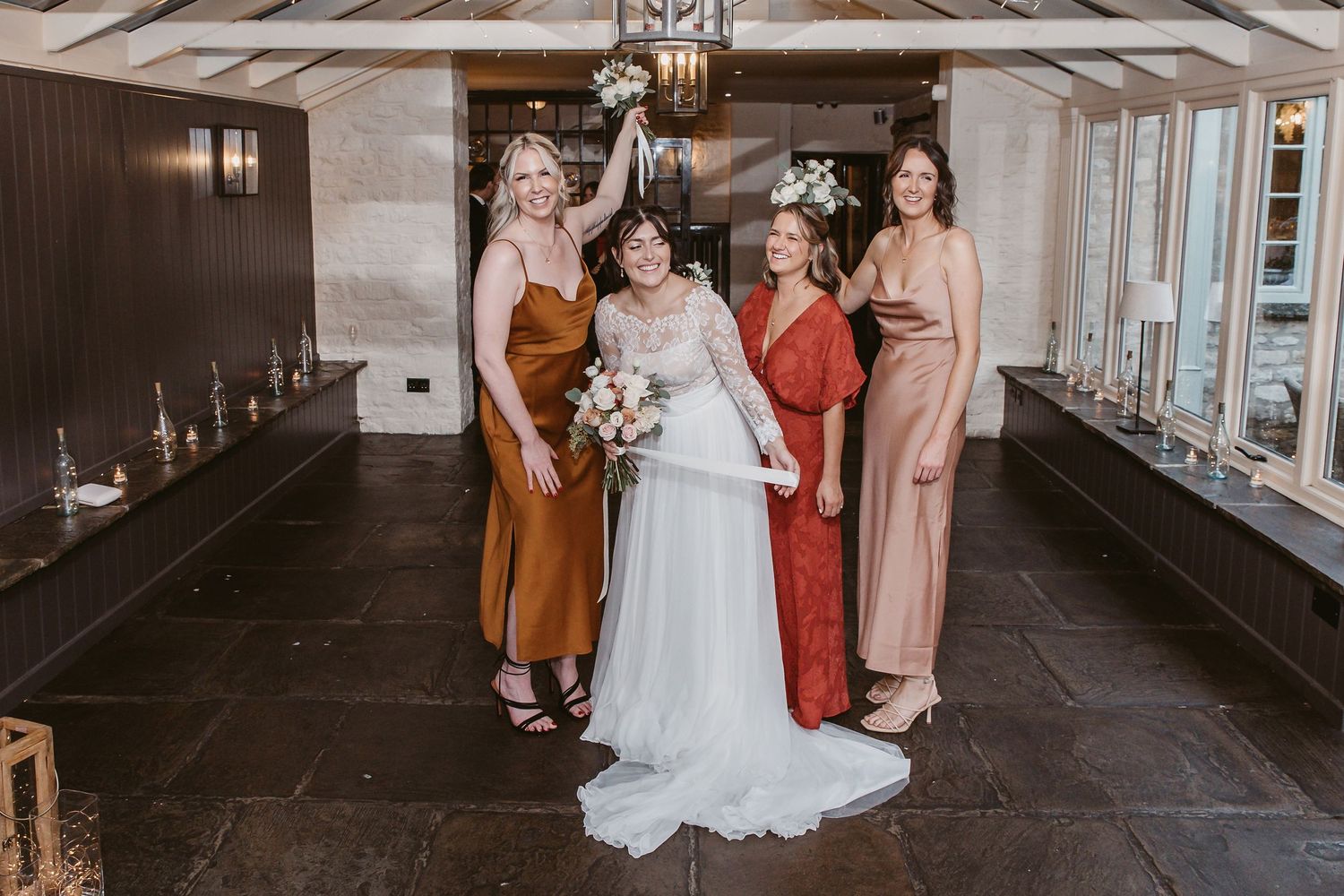 Bridal group wearing jewel-toned silk dresses pose together in elegant indoor setting.