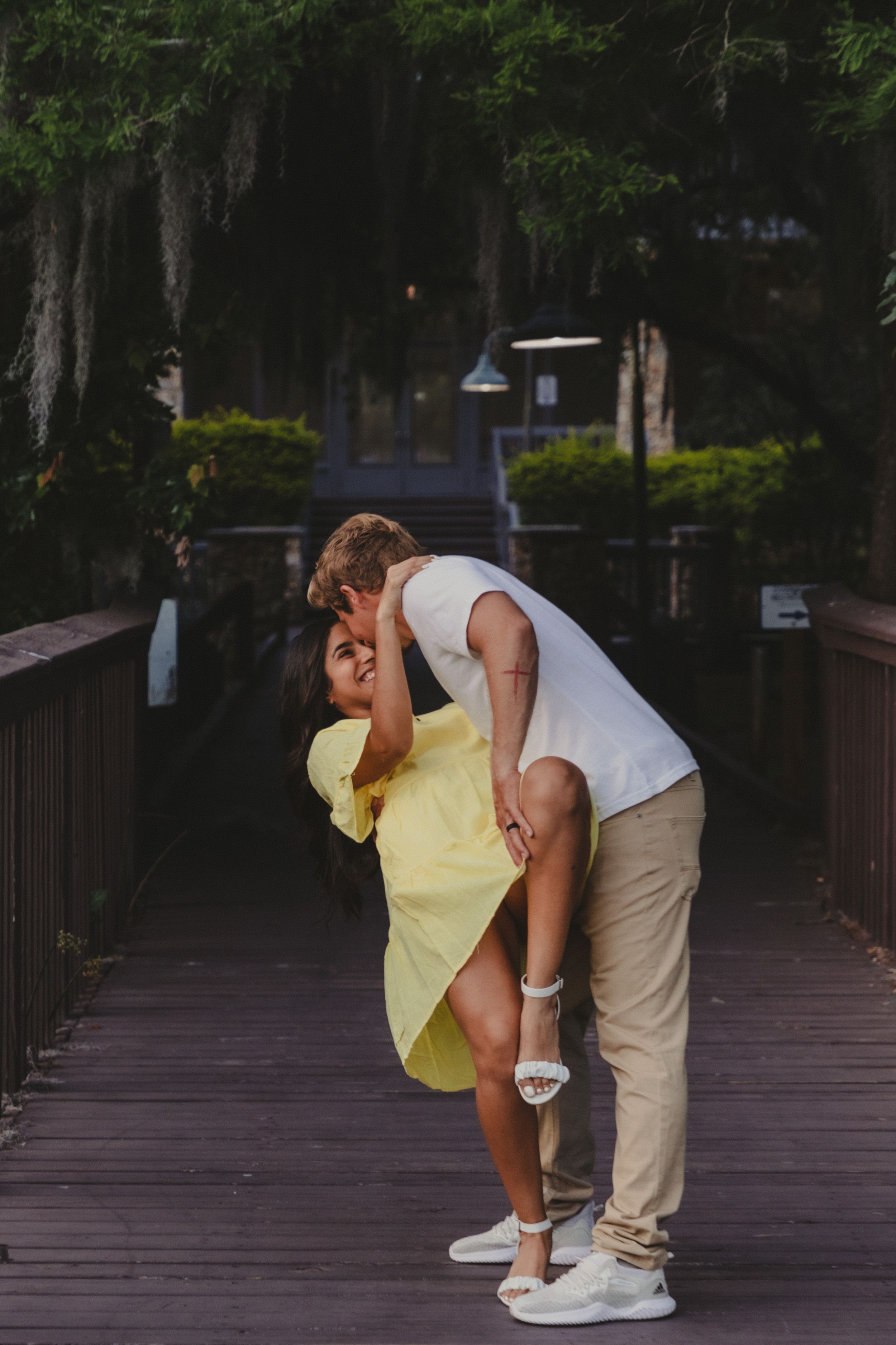 A couple shares a playful moment on a wooden boardwalk surrounded by trees.
