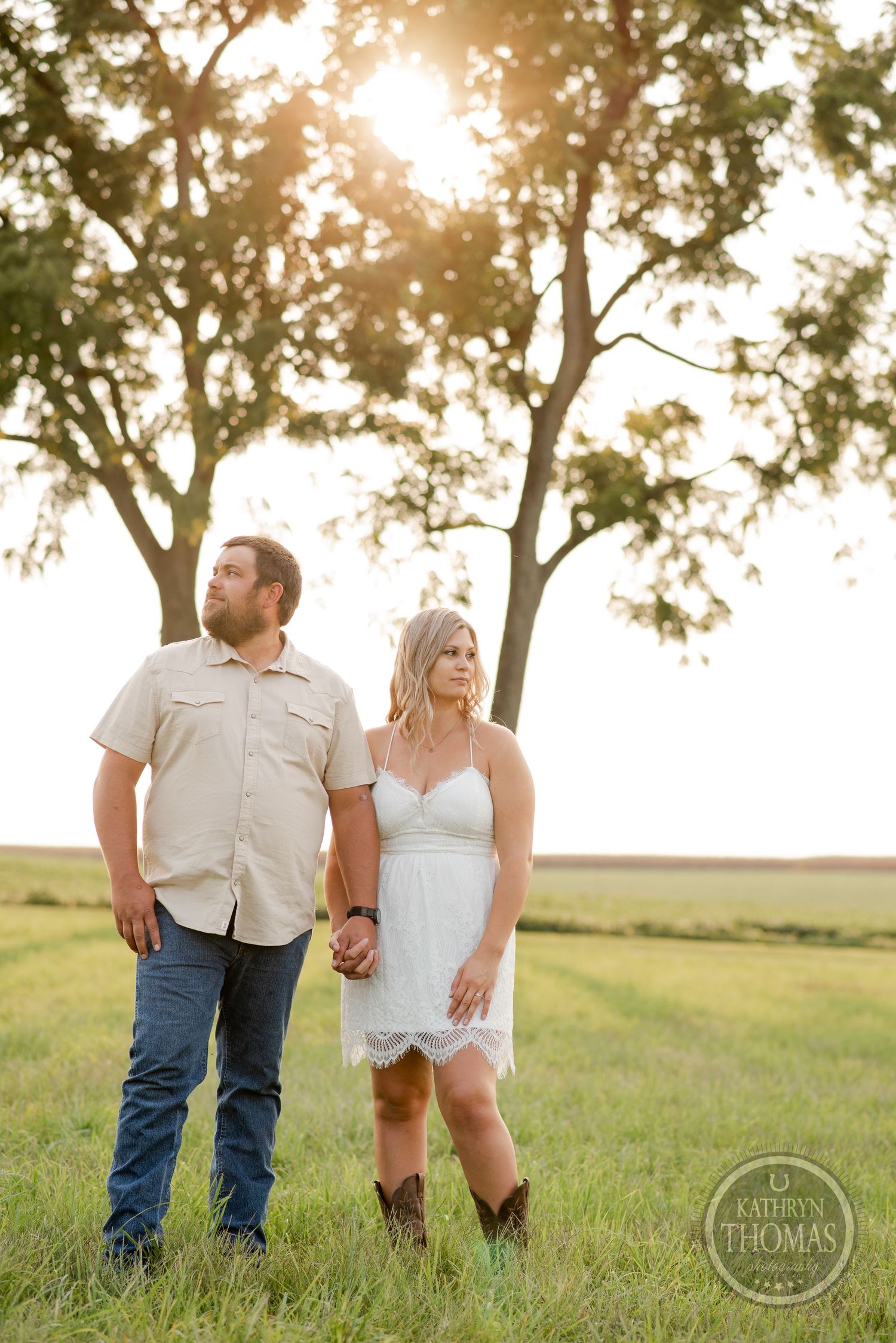 A couple stands together in a golden field during sunset with trees silhouetted in the background.