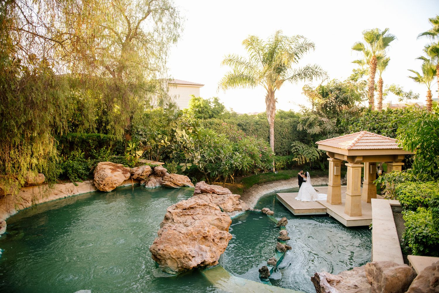 Tropical pool landscape with rock formations, palm trees and decorative gazebo at sunset.