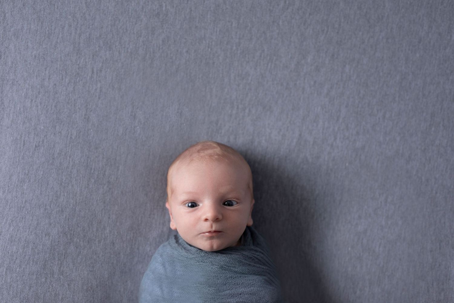 Baby in blue swaddle blanket lying against gray background looking at camera.