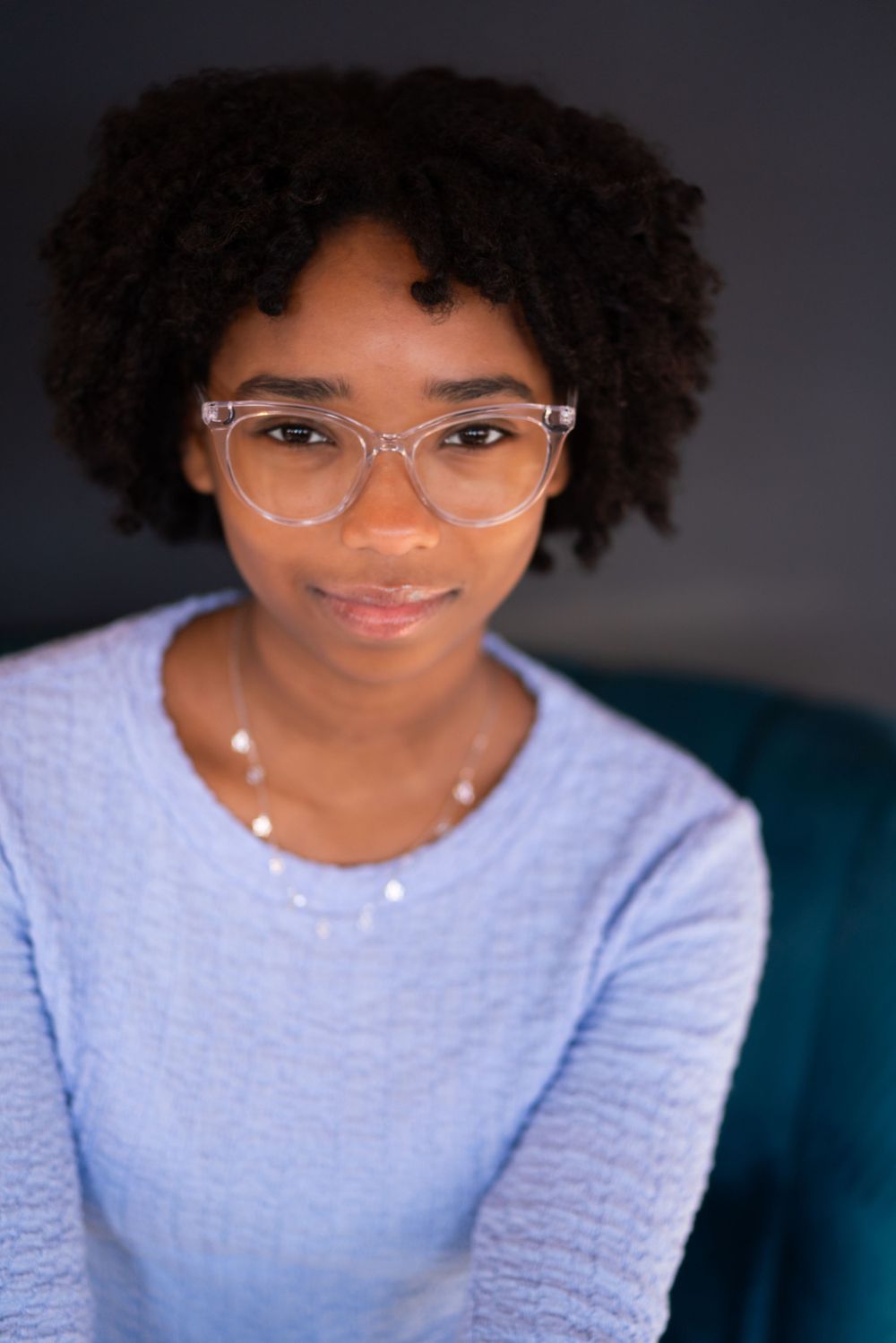 Professional headshot against dark background wearing a light blue sweater and clear glasses.