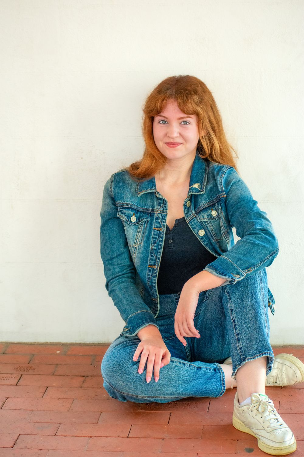 Person in denim jacket and jeans sits casually against white wall.