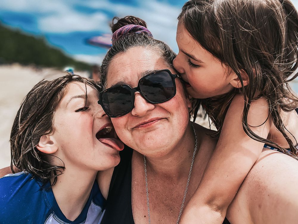 Sandwiched between her two kids on the beach, a mom smiles as she is kissed on each cheek.