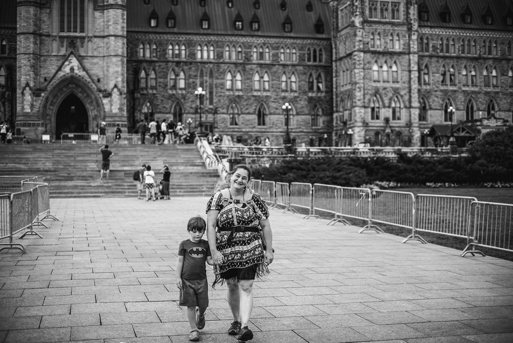 With the Canadian Parliament buildings in the background, a woman walks toward the camera with her young son beside her.