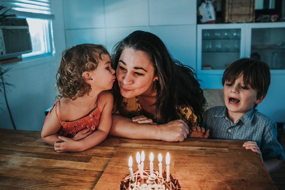 A cake is filled with lit candles as the birthday girl (mom) gets a kiss from her daughter as her son looks on.