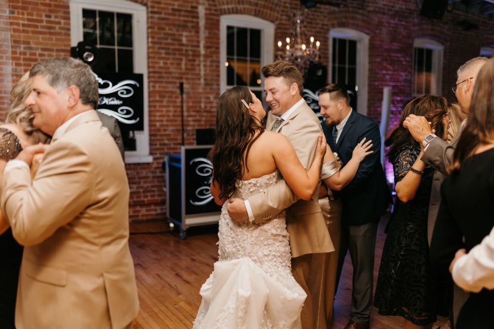 Bride in lace gown dancing with groom at lively wedding reception near brick wall backdrop.