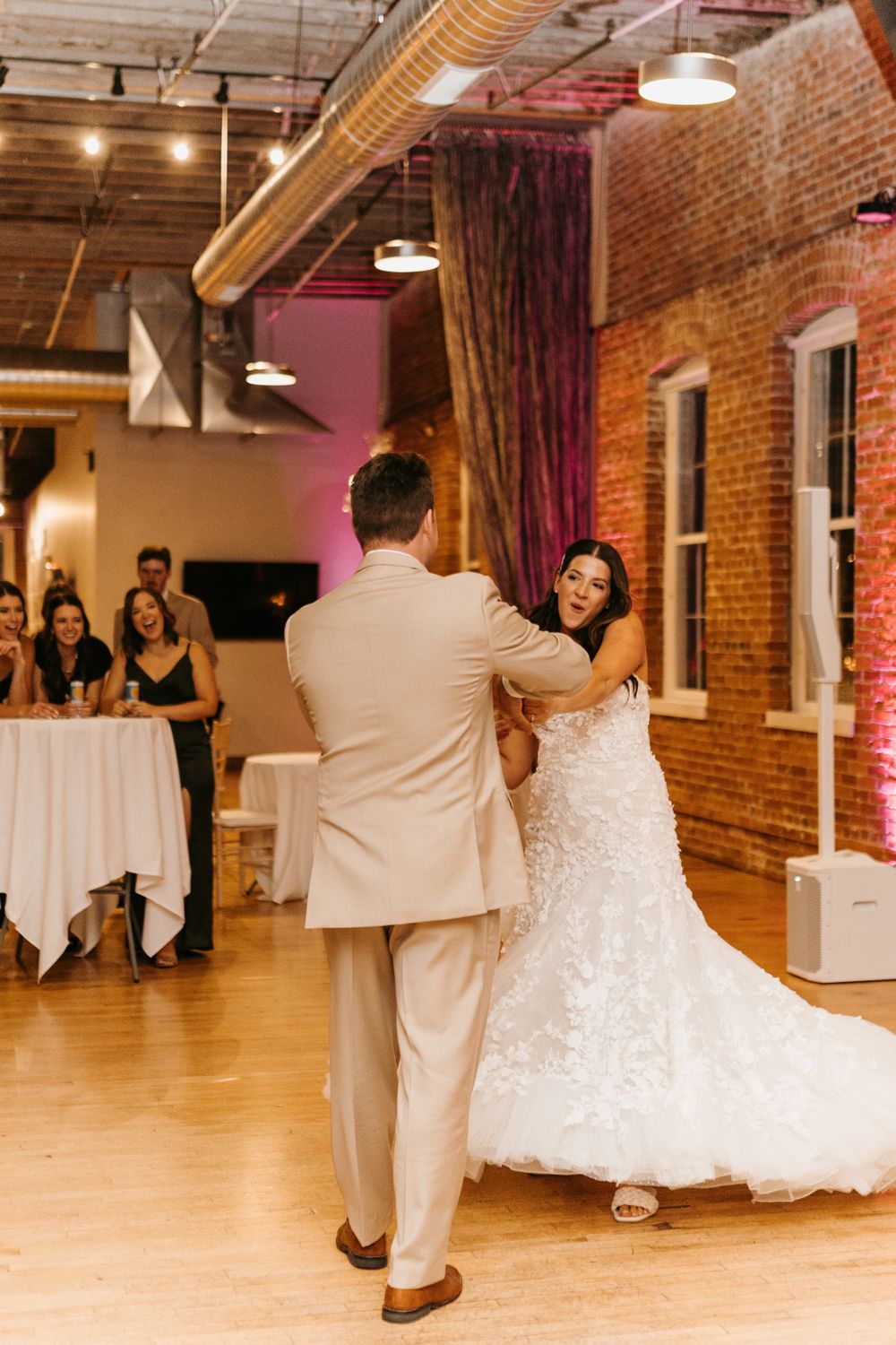 Bride in white lace gown twirls during first dance with groom at rustic brick-walled wedding venue.