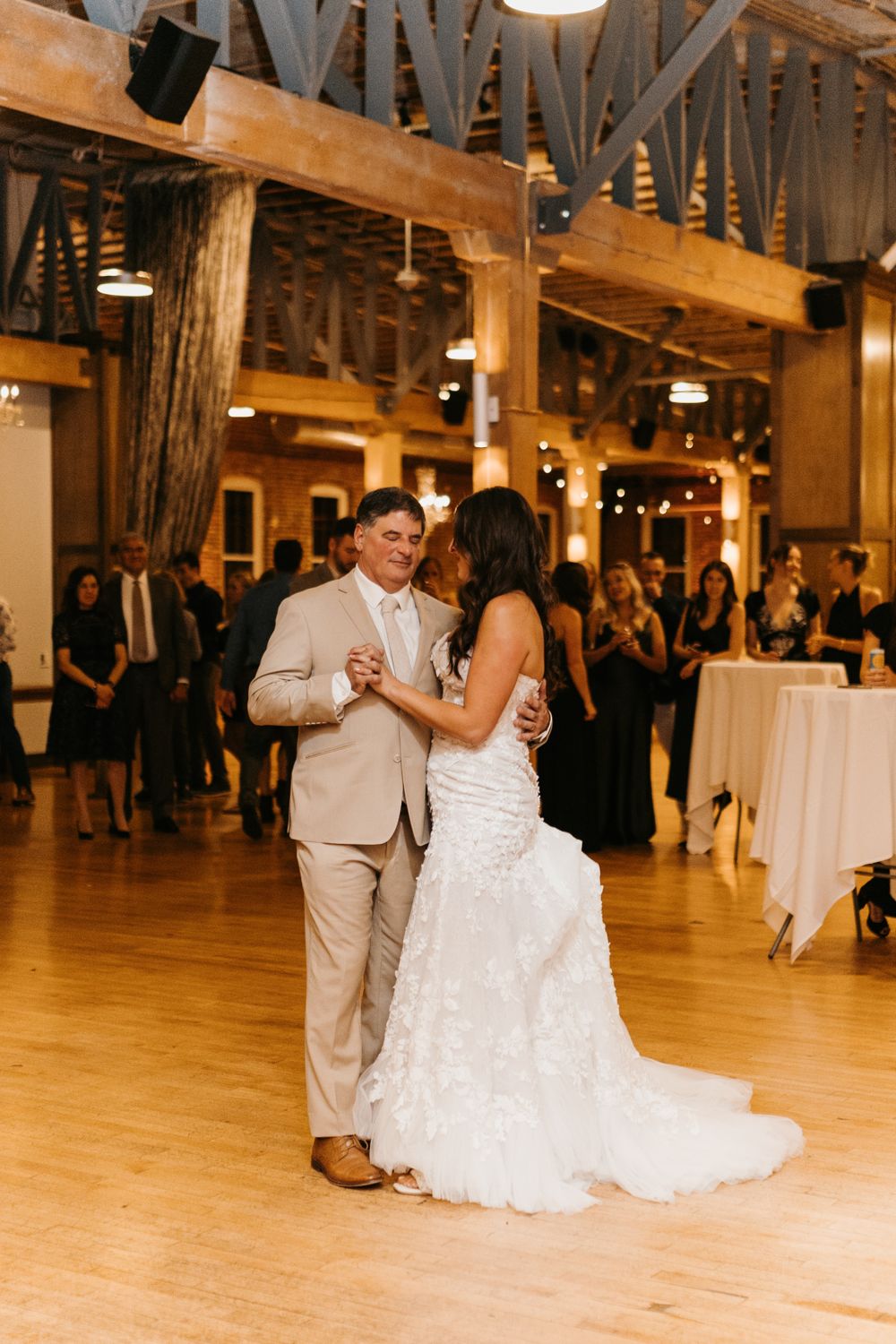 Bride and groom share elegant first dance at warmly lit rustic wedding reception with exposed wood beams.