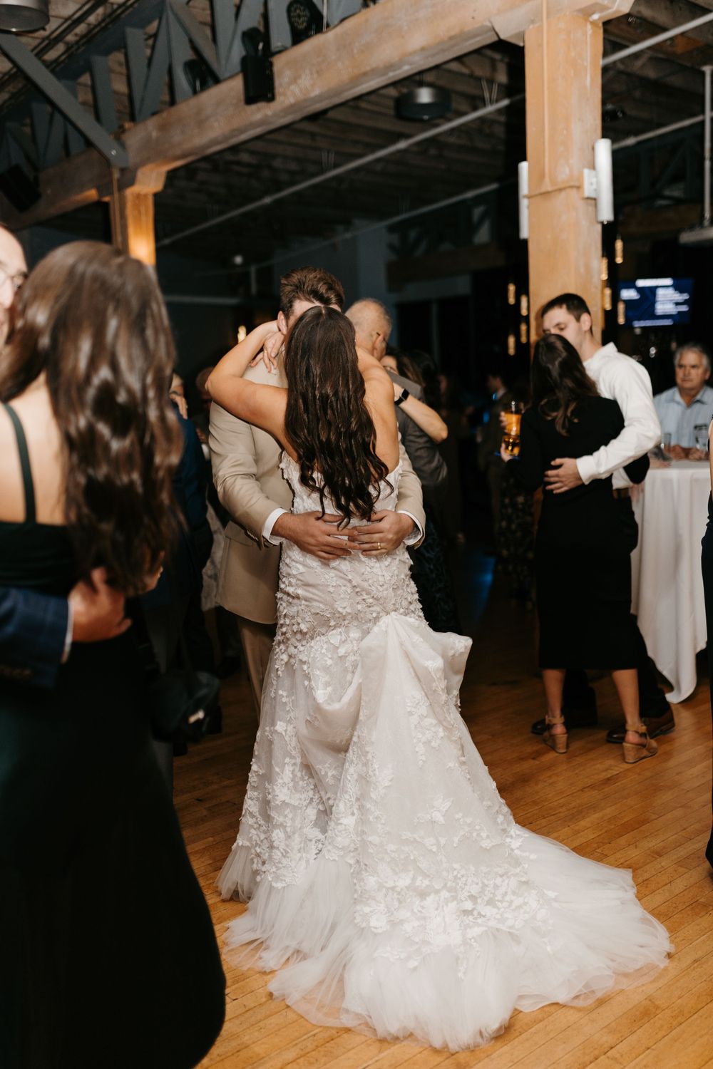 Bride in floral lace wedding gown embracing groom on crowded dance floor at wedding reception.