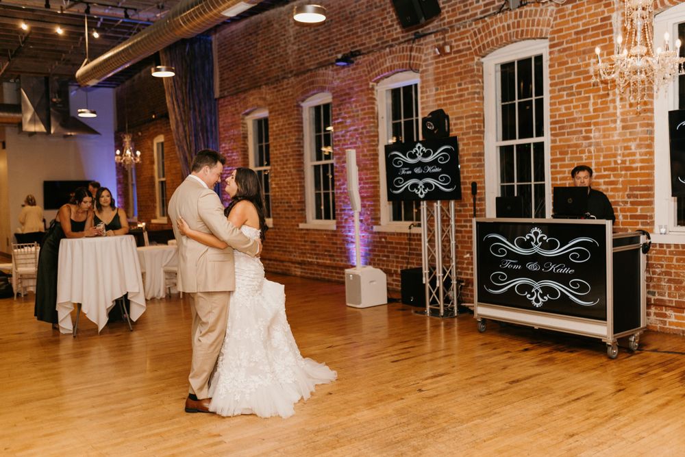 Bride and groom share first dance at elegant indoor wedding reception with DJ booth and brick wall backdrop.