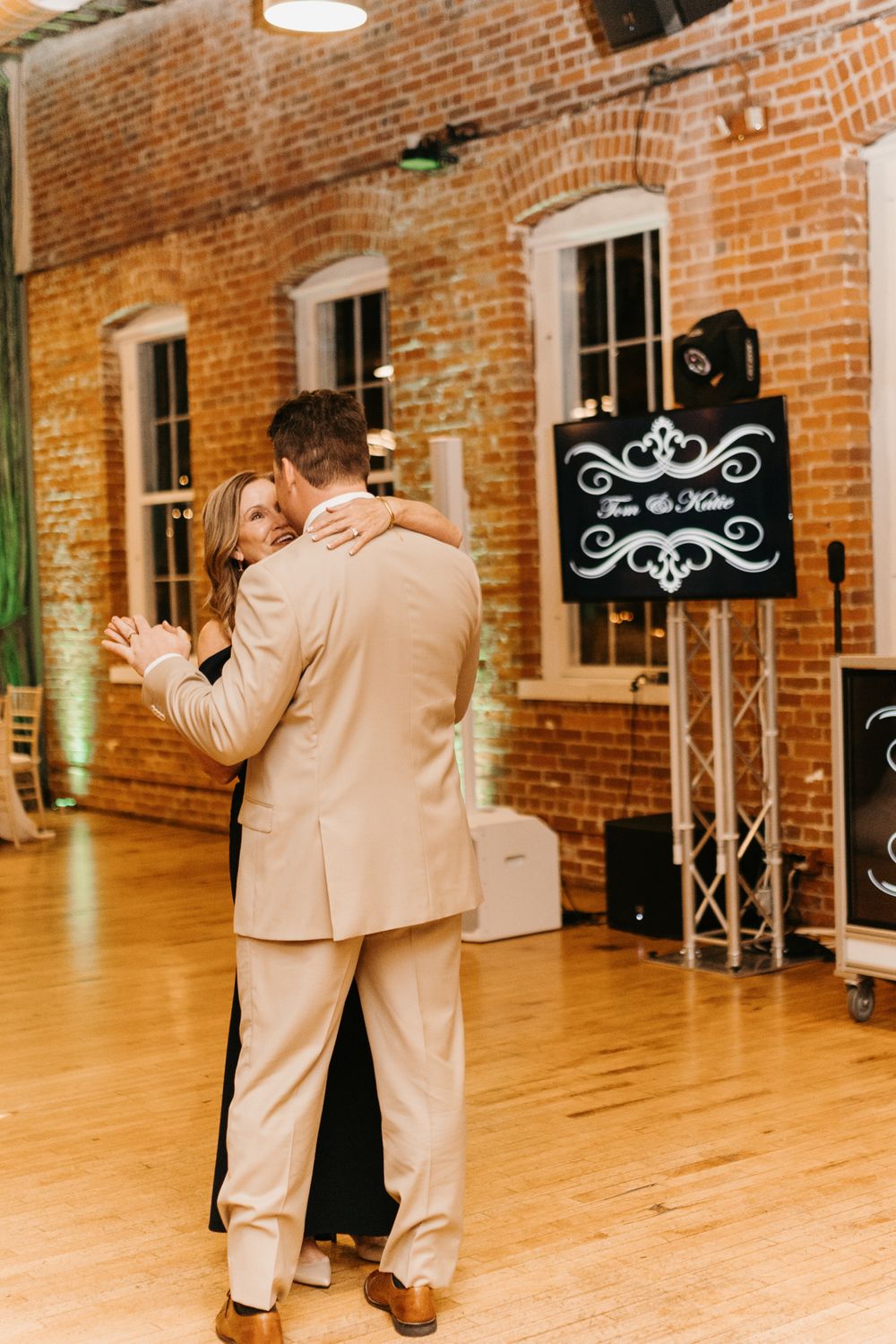 Groom dances with woman in black dress near personalized DJ booth at elegant wedding reception.