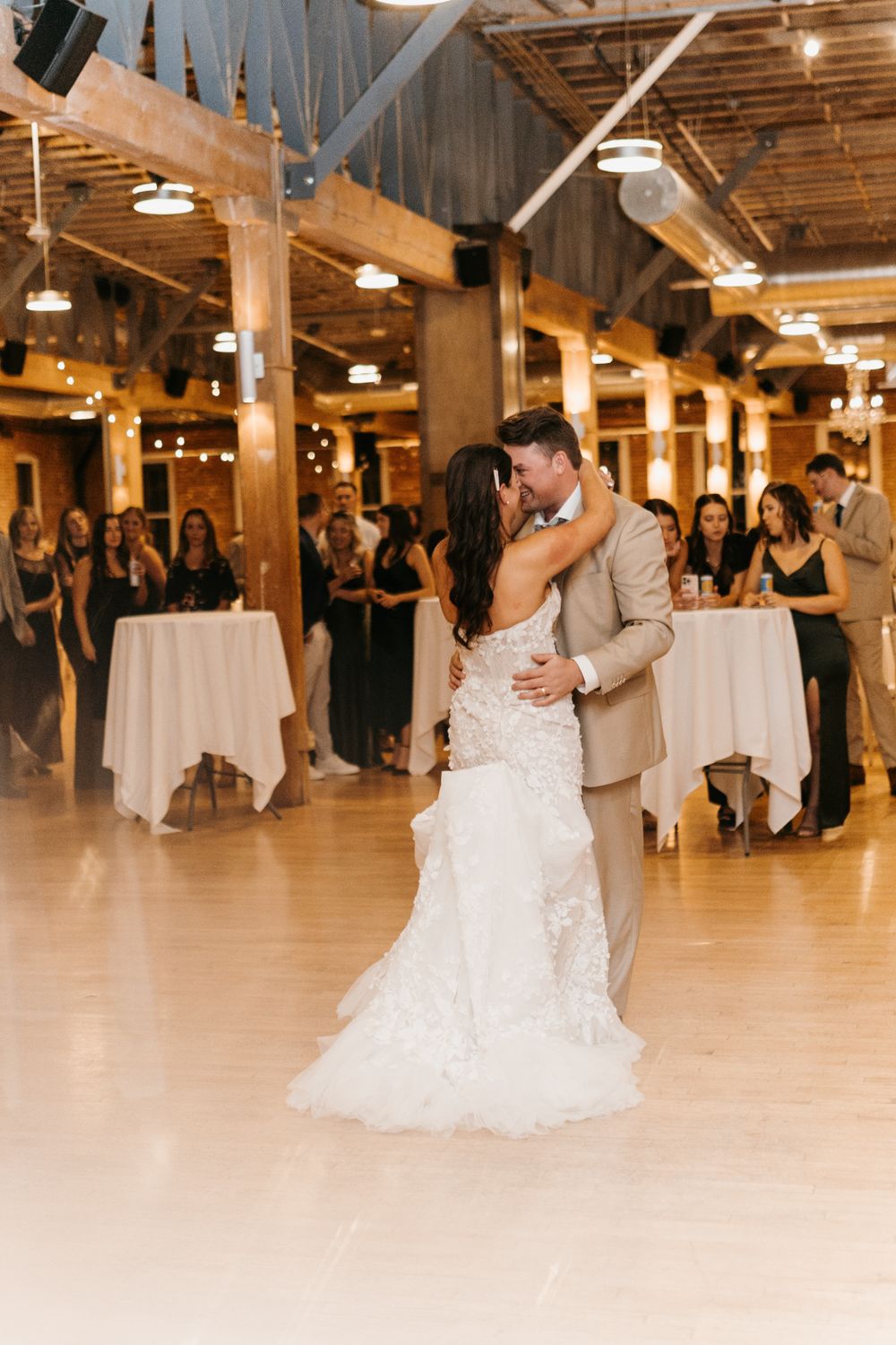 Couple shares emotional first dance surrounded by wedding guests at rustic industrial reception venue.