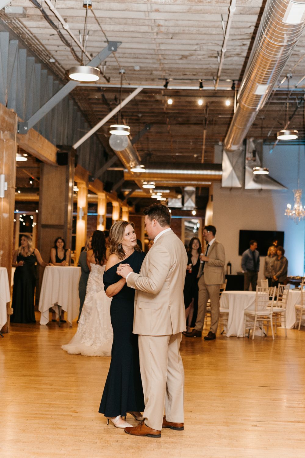 Groom in tan suit dancing with woman in black gown at an industrial-chic wedding reception.