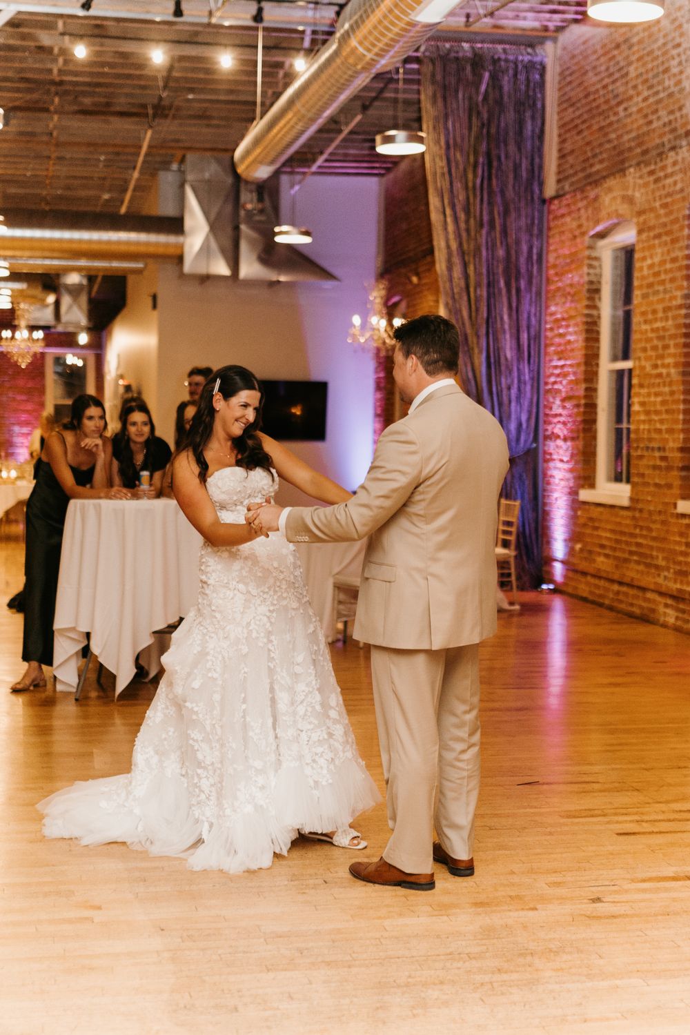 Smiling bride in floral lace gown holds hands with groom in tan suit during first dance at reception.