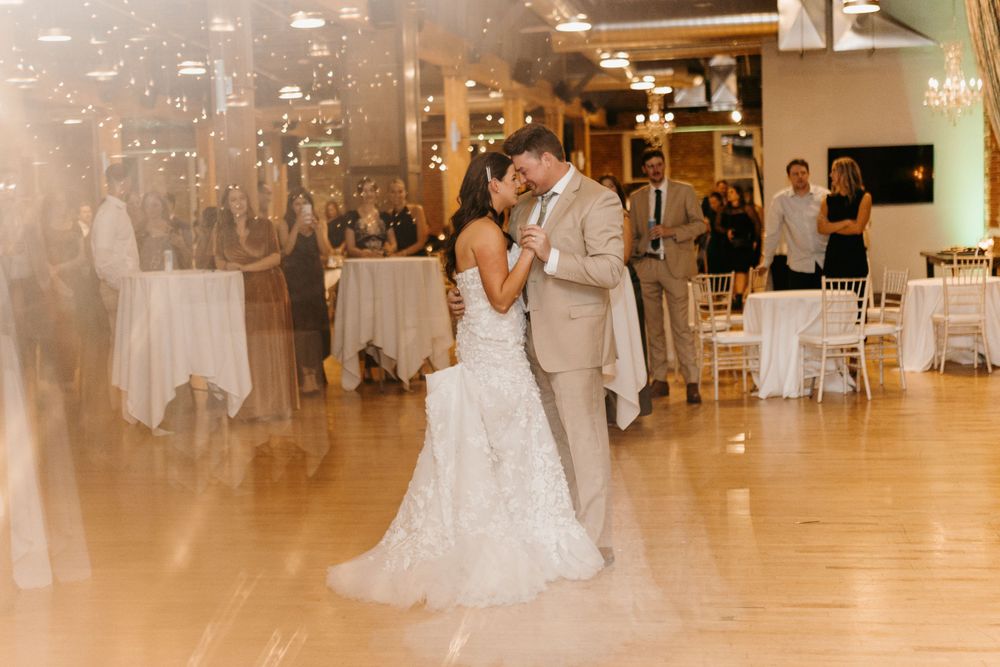 Bride in lace gown and groom in tan suit share romantic first dance at warmly lit wedding reception.