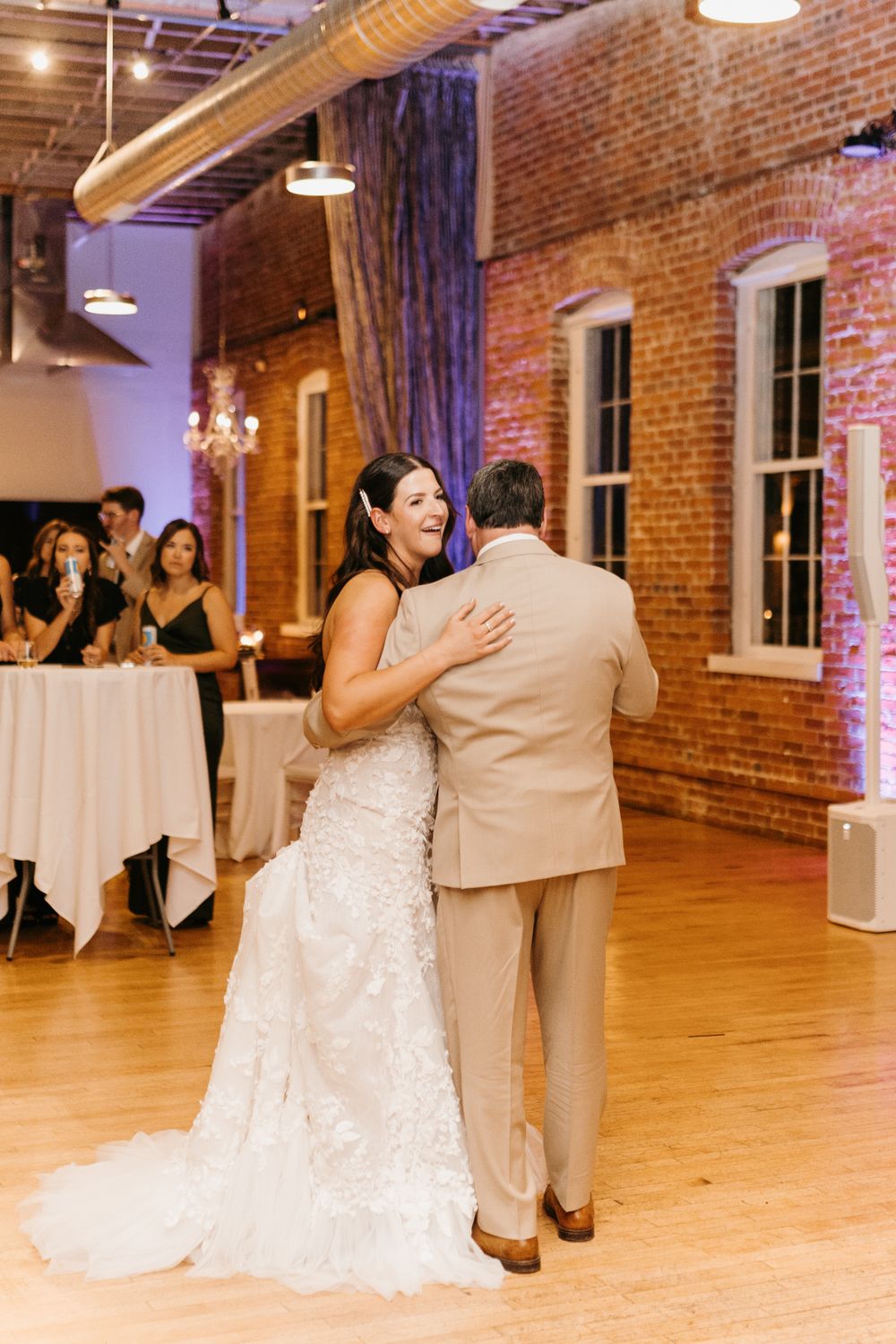 Laughing bride in lace gown embraces groom during first dance at brick-walled wedding reception.