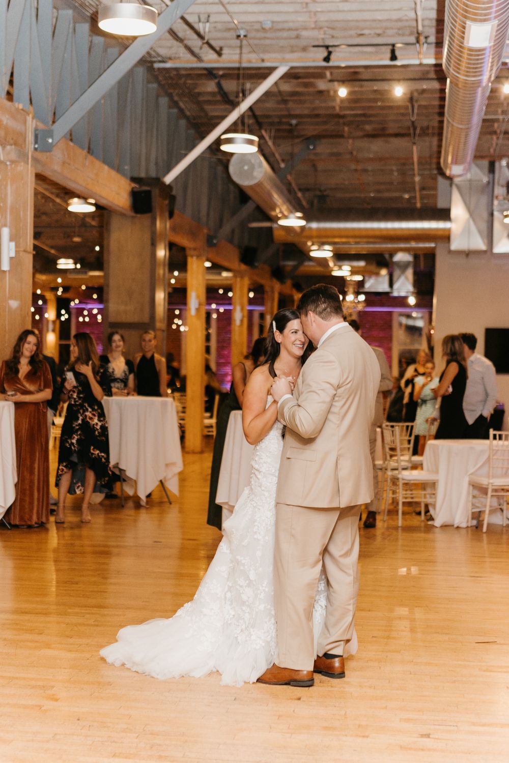 Newlyweds share an intimate first dance on wooden floor at modern industrial wedding venue with guests watching.