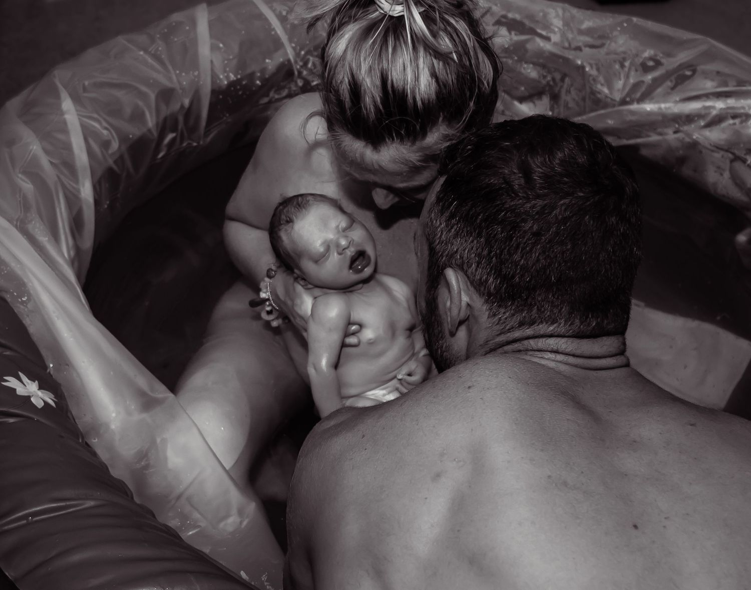 Black and white image of a mother sitting in a birthing tub holding her newborn baby as her husband looks.