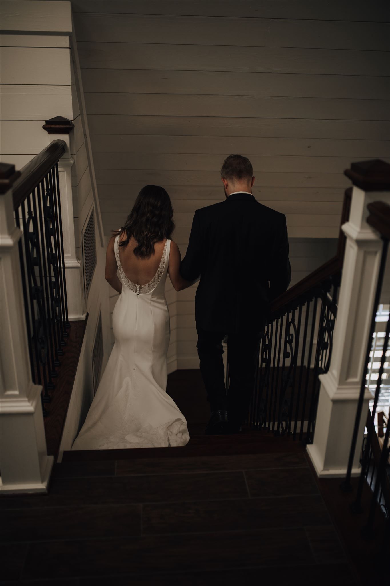 Bride and groom descending elegant staircase with iron railings in dramatic lighting.