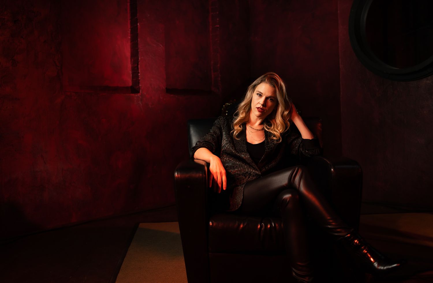 A woman sits in a chair wearing all black. The scene is dramatically lit in red.