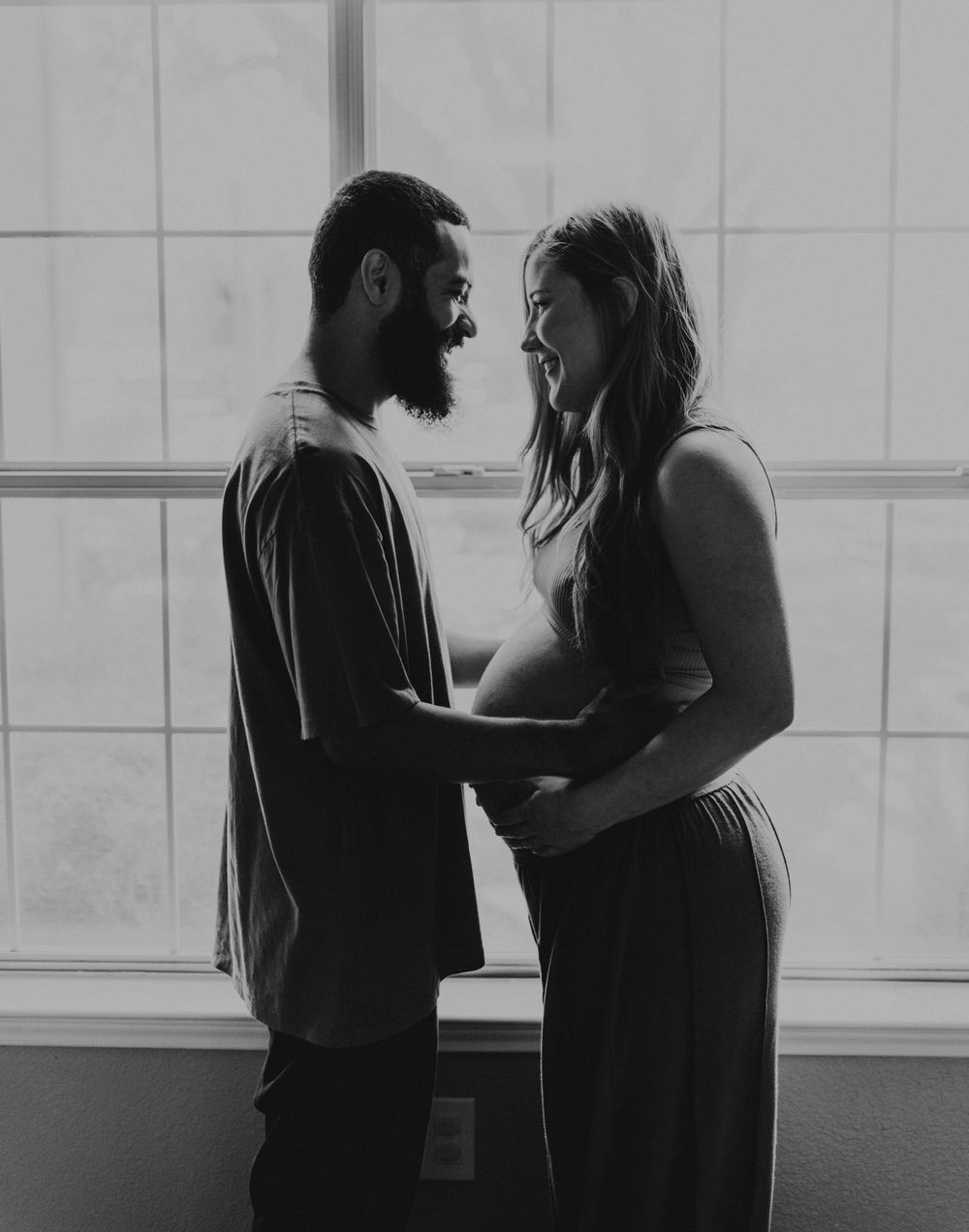 Black and white maternity photo showing couple's intimate moment by window.