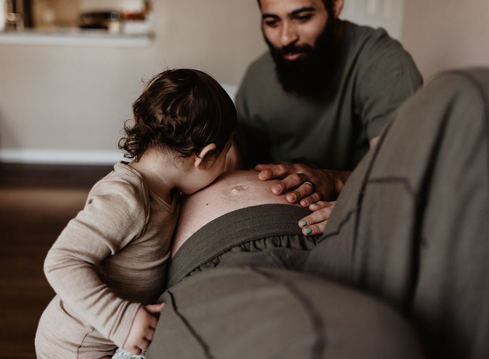 Toddler and adult hands touch a pregnant belly on a cozy couch.