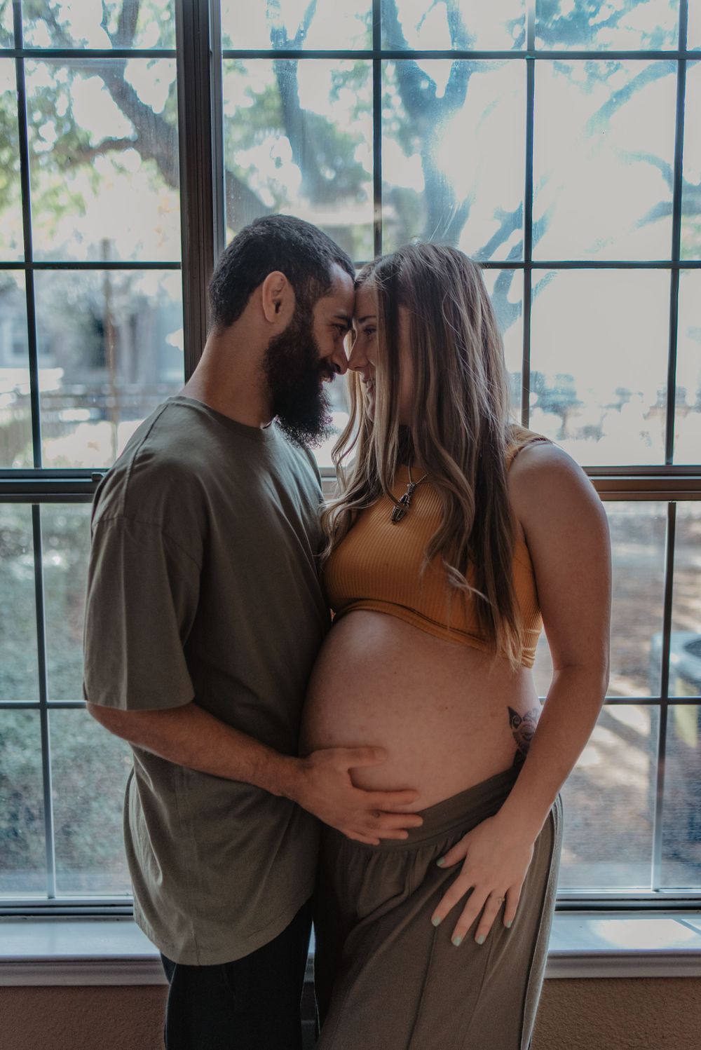 Expectant couple shares intimate moment by large window during maternity photoshoot.