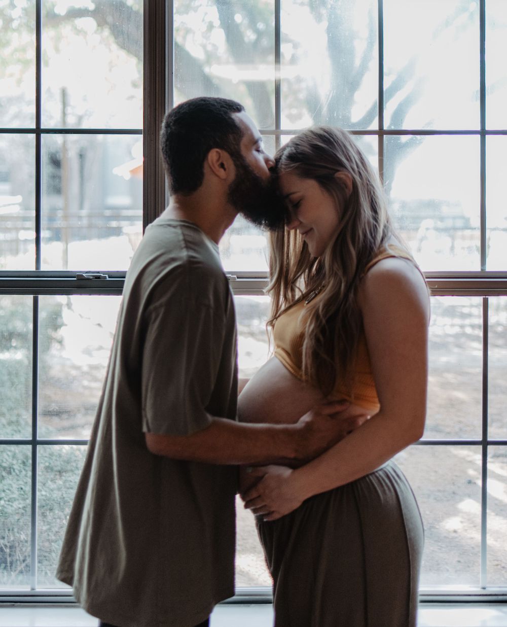 Intimate maternity photo of couple embracing by window with natural light.