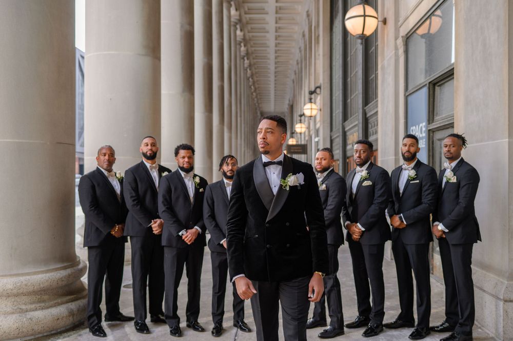 Groomsmen in black suits pose between classical columns in an elegant hallway.