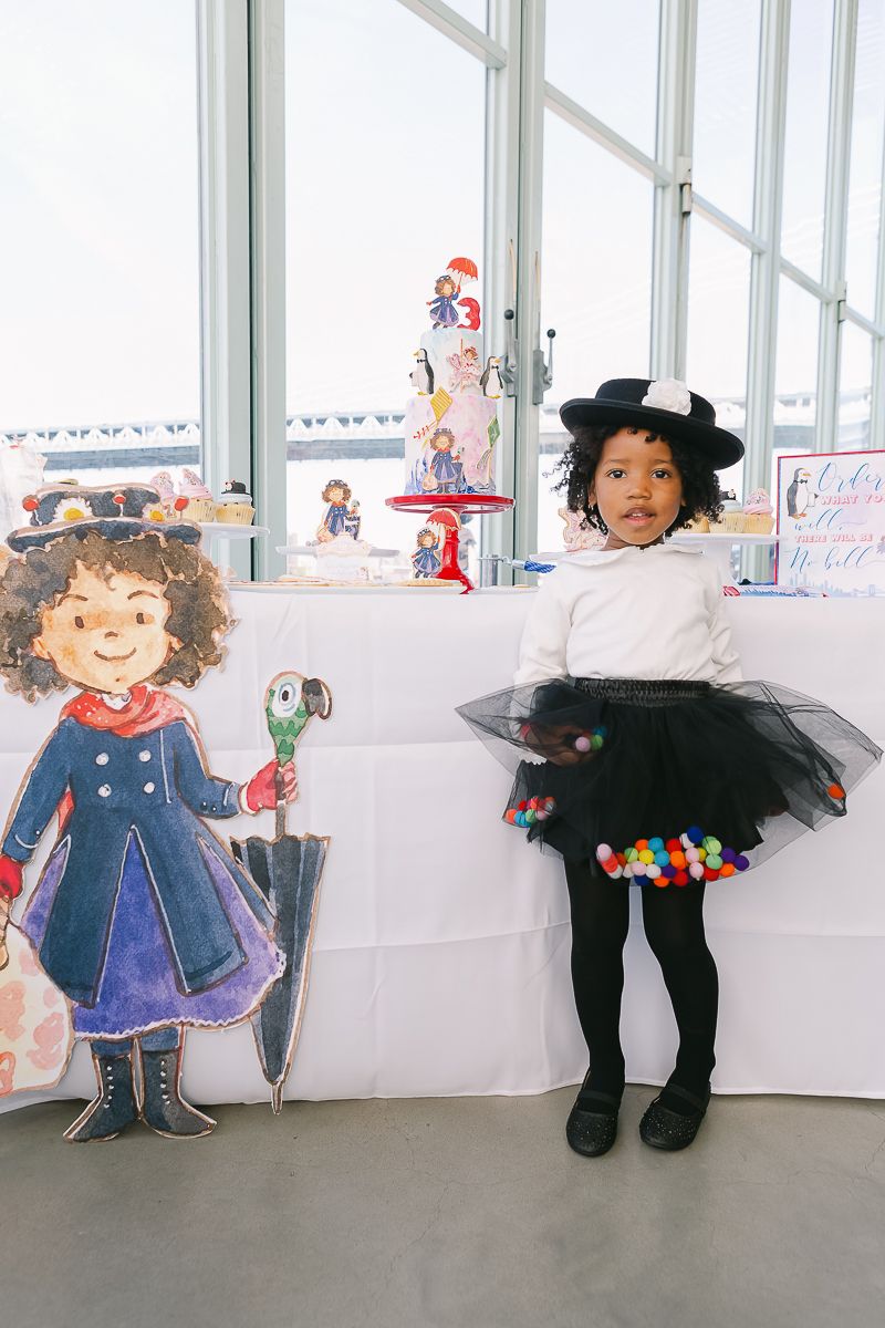 a birthday girl wearing a black hat and tutu, and white shirt stands next to her Mary Poppins birthday party decor