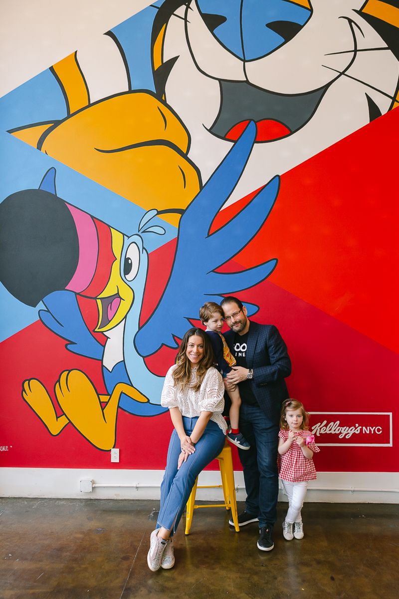 a couple with their toddler son and daughter stand in front of a Kelloggs' mural in Manhattan during a birthday party