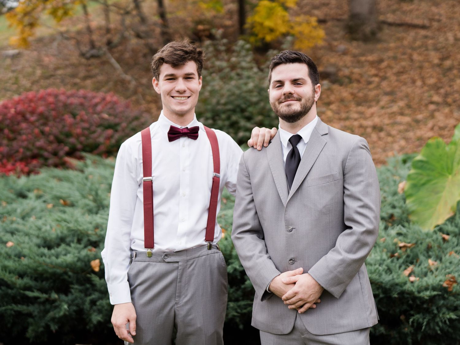 Stylish groom and best man in burgundy bow tie and suspenders pose in autumn garden setting.