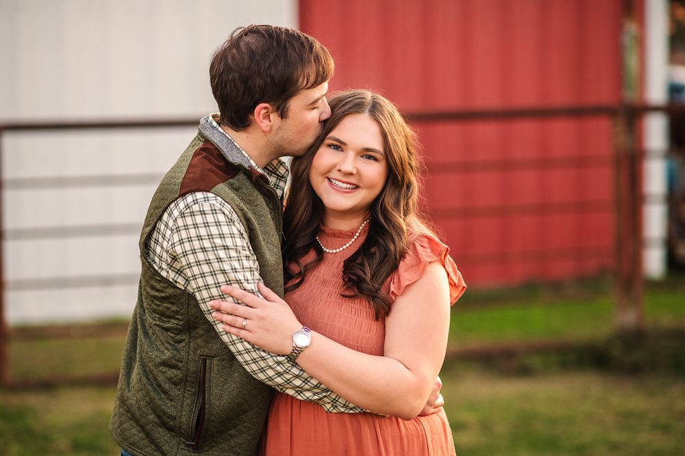 Couple embracing outdoors near red barn, man kissing woman's cheek as she smiles warmly.