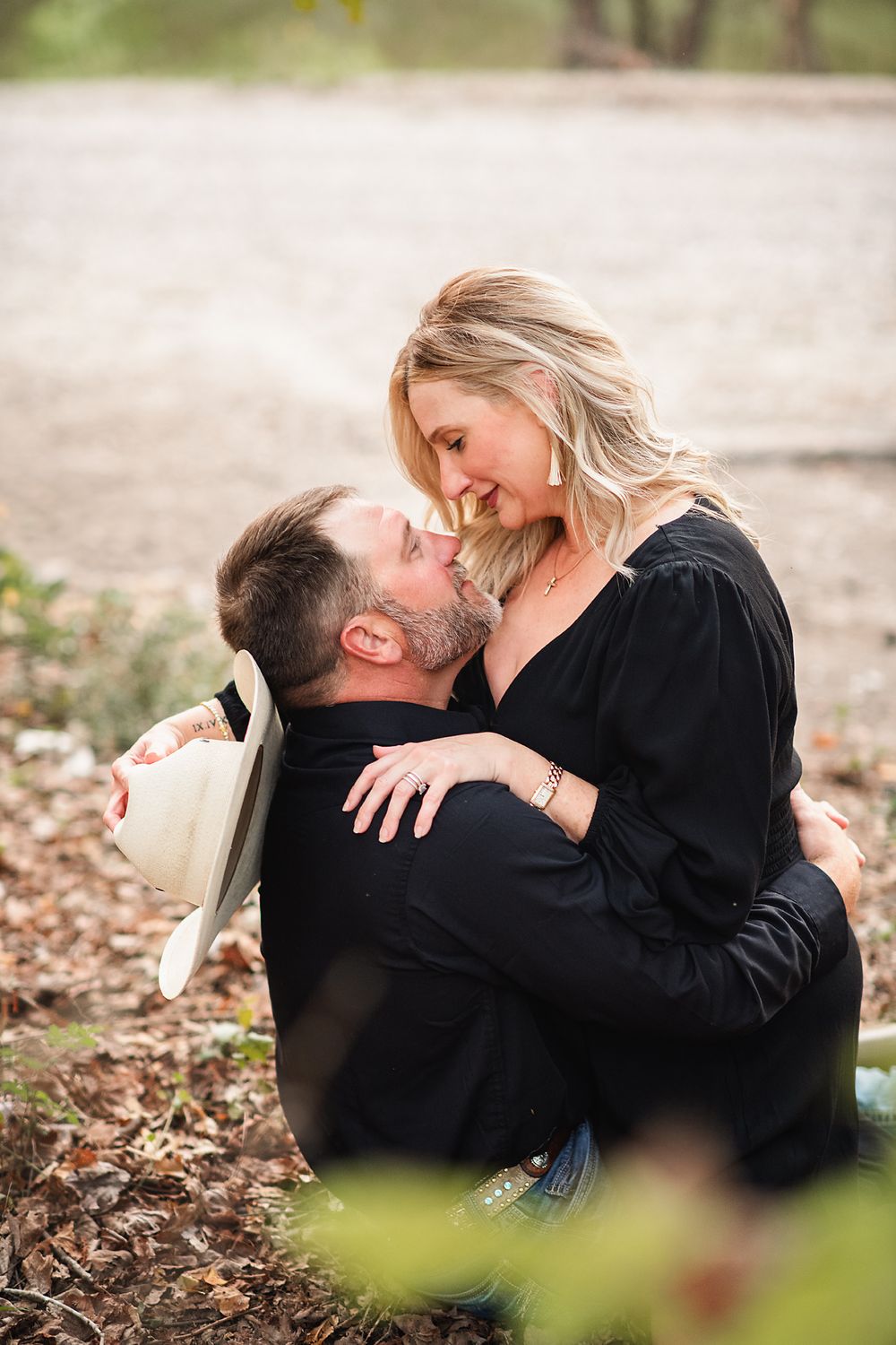 Couple sharing a romantic moment outdoors, man kneeling and kissing woman in black dress among autumn leaves.