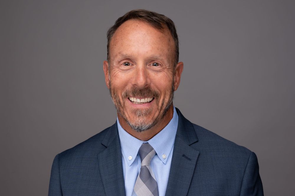 Professional headshot of smiling businessman in navy suit and blue tie against gray background.