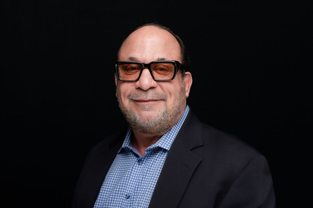 Professional headshot of a person wearing glasses and a suit jacket against a black background.