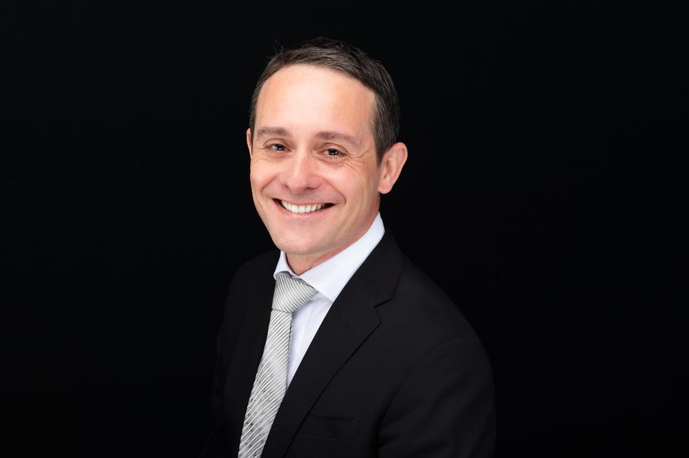 Professional headshot portrait against black background showing business attire and warm smile.
