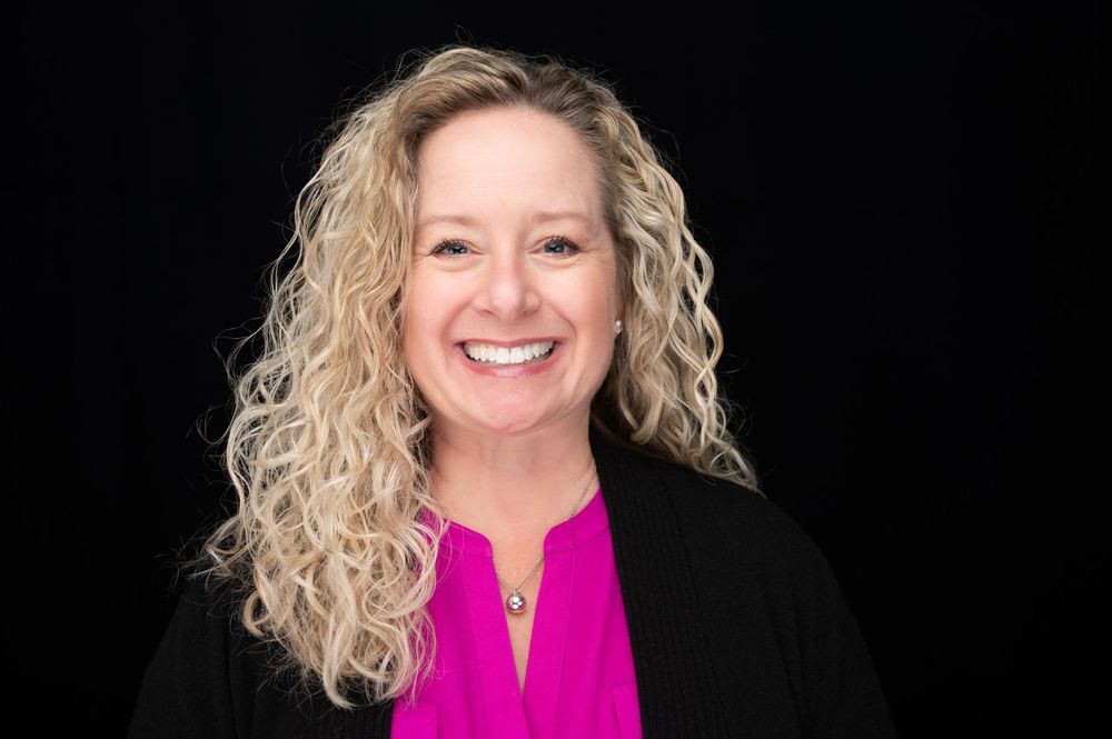 Professional portrait of woman wearing pink top and black blazer against dark background.