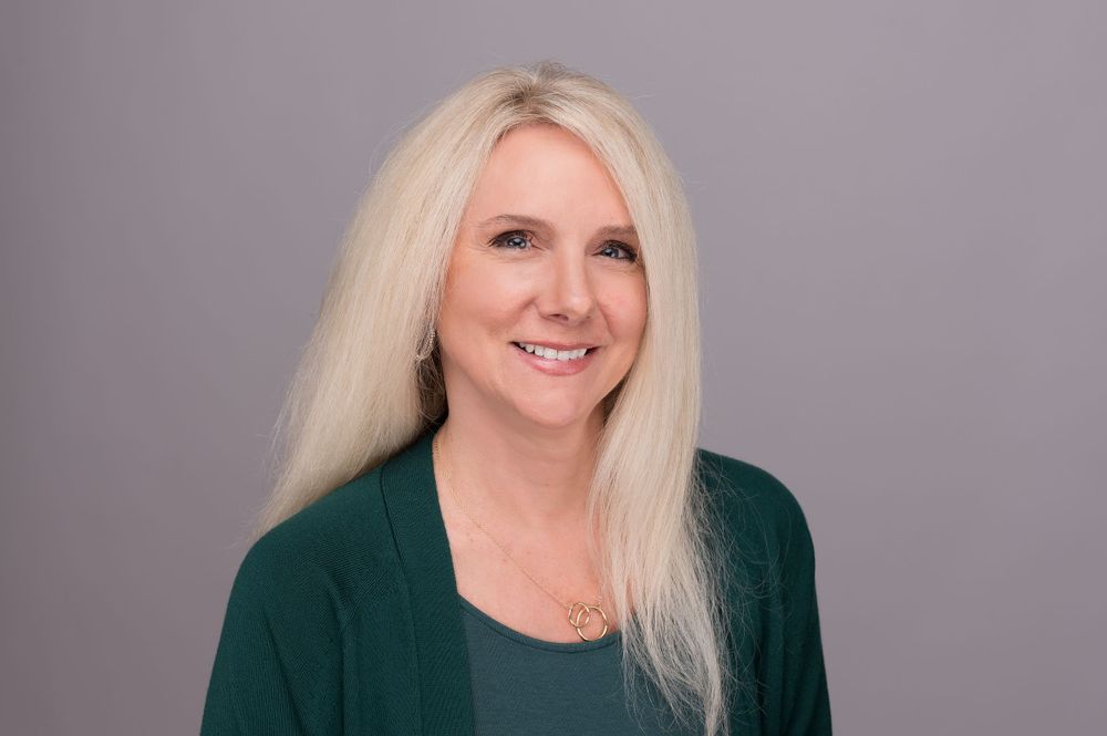 Professional headshot of woman with platinum hair wearing green top against gray background.