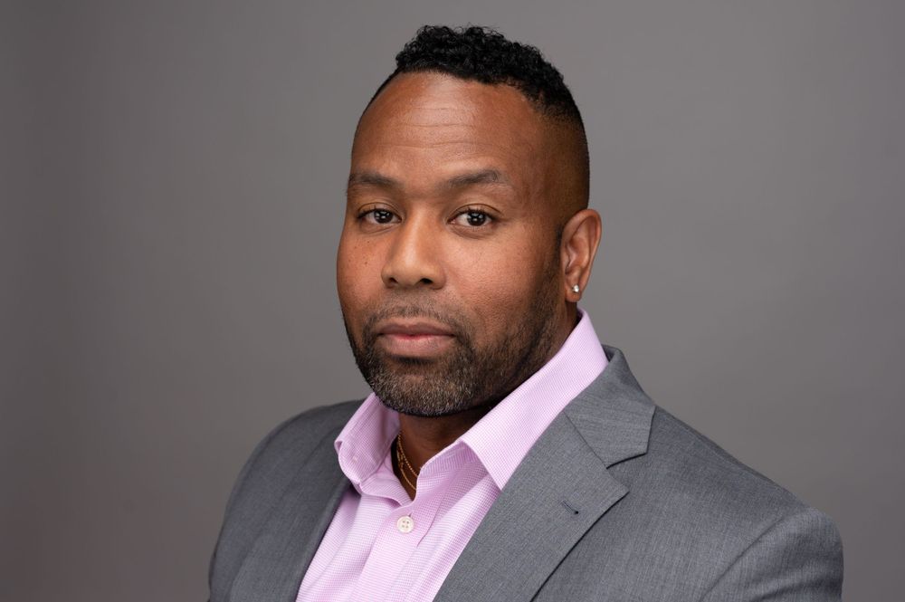 Professional headshot of person wearing grey suit and pink shirt against grey background.