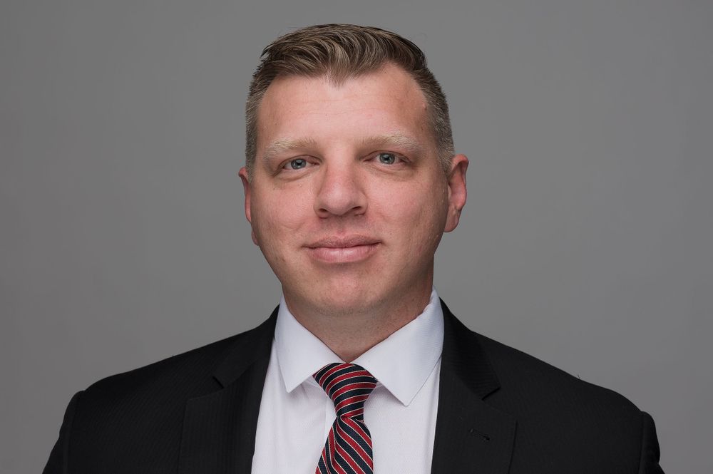 Professional headshot of businessman in dark suit and red tie against grey backdrop.