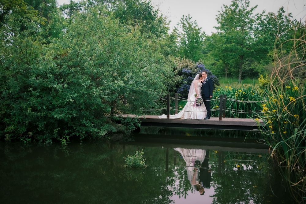 A couple shares a romantic moment on a wooden dock over a peaceful pond surrounded by lush greenery.