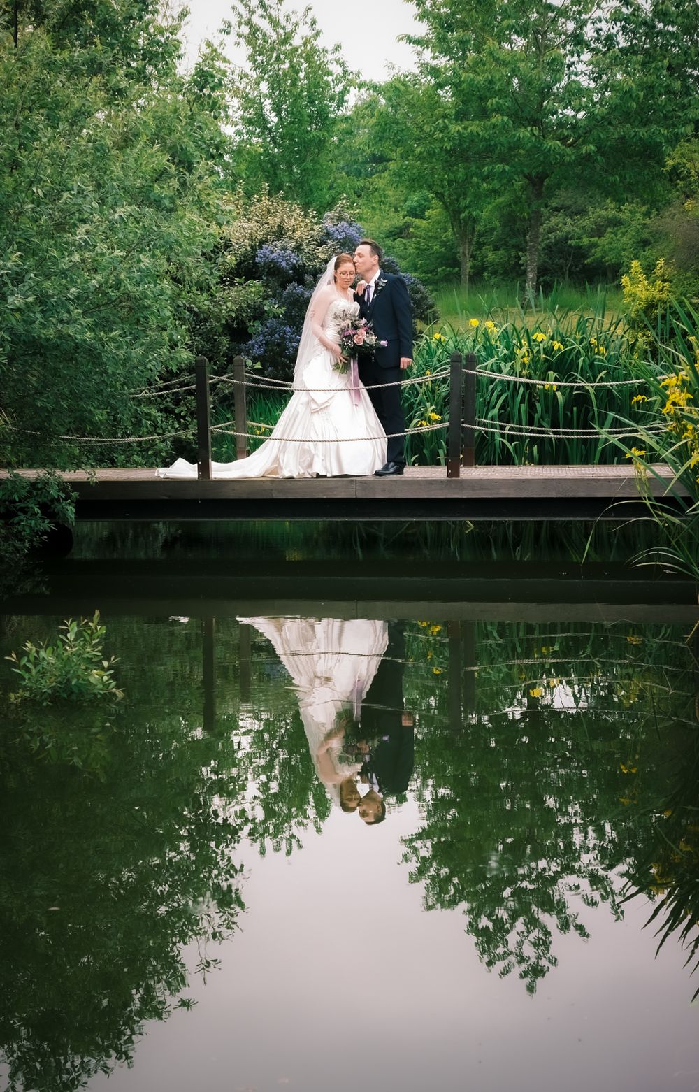 Wedding couple on wooden bridge with perfect reflection in pond surrounded by green foliage.